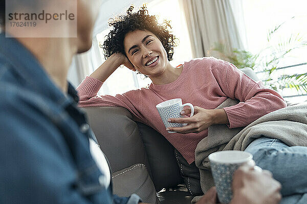 Glückliche junge Frau hält eine Kaffeetasse in der Hand und blickt ihre Freundin an  die zu Hause auf dem Sofa sitzt