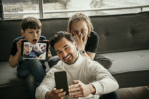 Glücklicher Mann  der mit seinem Smartphone ein Selfie macht  während seine Söhne zu Hause Grimassen schneiden