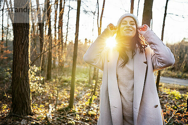 Happy woman in knit hat standing by trees in forest on sunny day