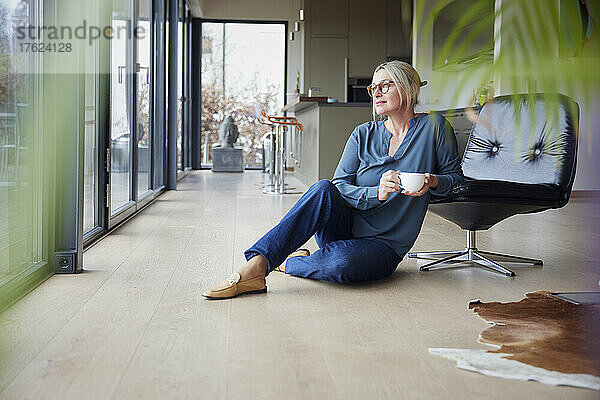 Blonde Frau mit Kaffeetasse sitzt zu Hause am Stuhl