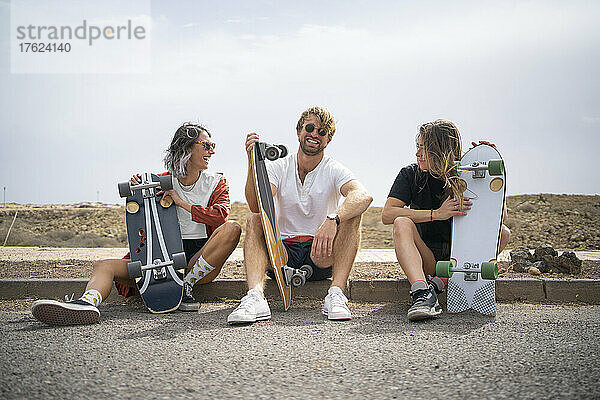 Fröhliche Freunde mit Skateboards sitzen an sonnigen Tagen am Straßenrand