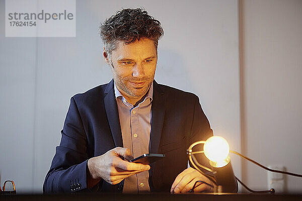 Geschäftsmann steuert Glühbirne mit Smartphone am Arbeitsplatz