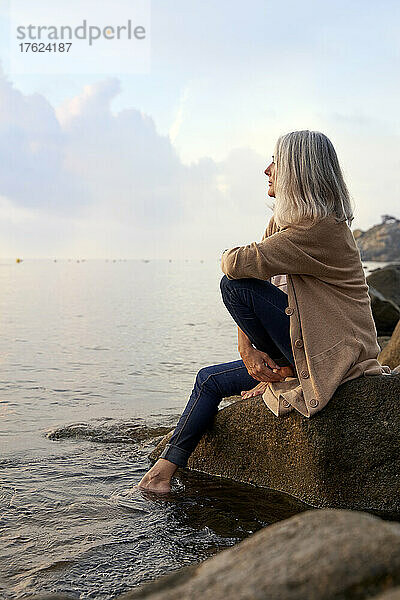 Frau sitzt auf einem Felsen und blickt aufs Meer