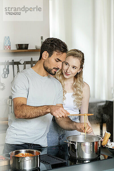 Lächelnde blonde Frau schaut ihrem Freund zu  der in der Küche Pasta macht