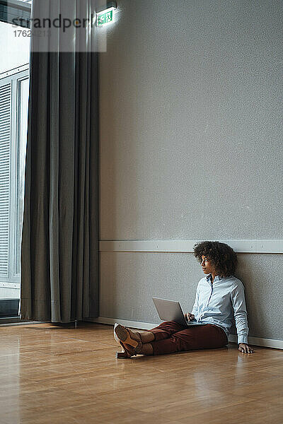 Nachdenkliche Geschäftsfrau mit Laptop sitzt auf dem Boden im Büro
