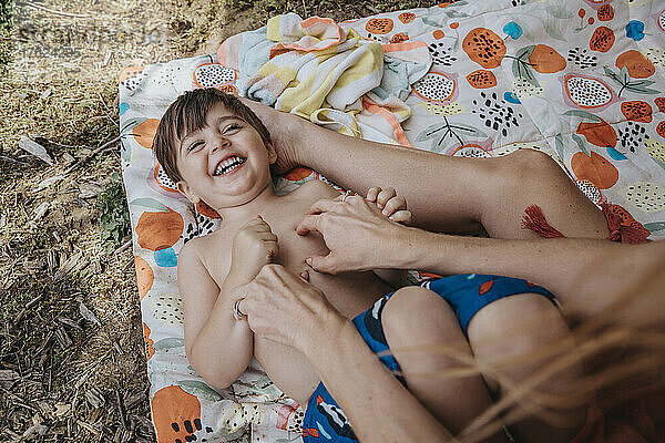 Mother tickling son lying on blanket at lakeshore