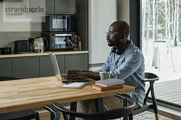 Geschäftsmann benutzt Laptop am Esstisch und sitzt zu Hause auf einem Stuhl in der Küche