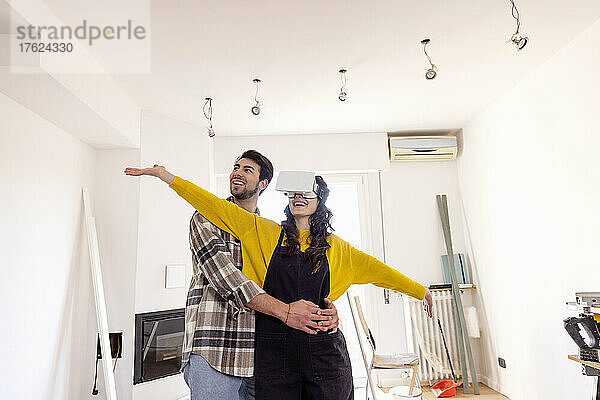 Man embracing happy girlfriend wearing VR glasses in new living room