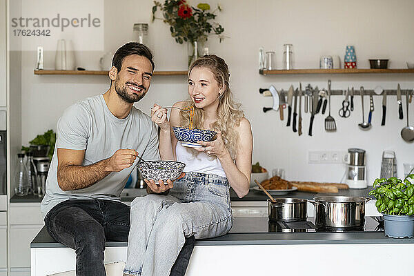 Lächelnde junge Frau und Mann essen Spaghetti und sitzen zu Hause auf der Kücheninsel
