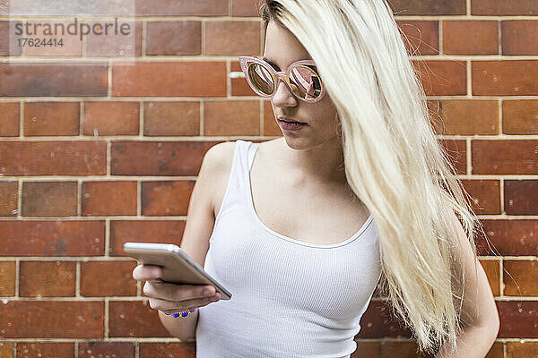 Blond teenage girl surfing net through smart phone in front of brick wall