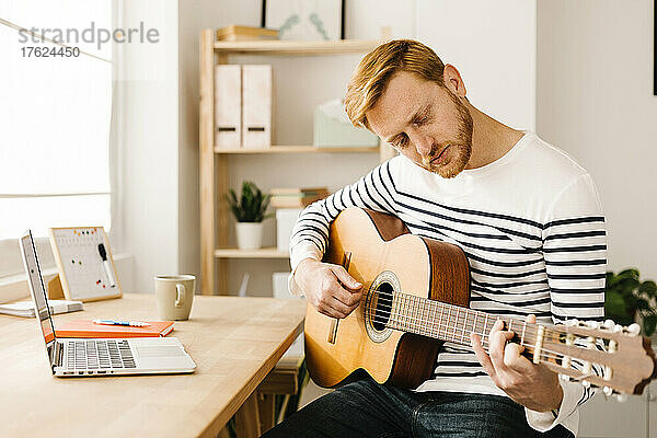 Junger Mann übt Gitarre und sitzt am Tisch im Wohnzimmer