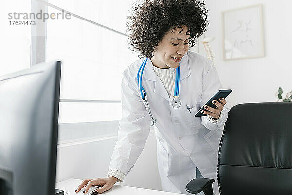 Lächelnder Arzt mit lockigem Haar  der in der Klinik sein Smartphone benutzt