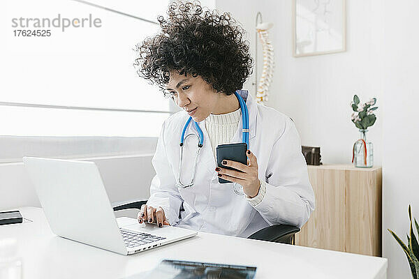 Junger Arzt hält Smartphone und Laptop in der Klinik in der Hand