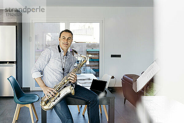 Lächelnder Musiker mit Saxophon sitzt zu Hause am Tisch
