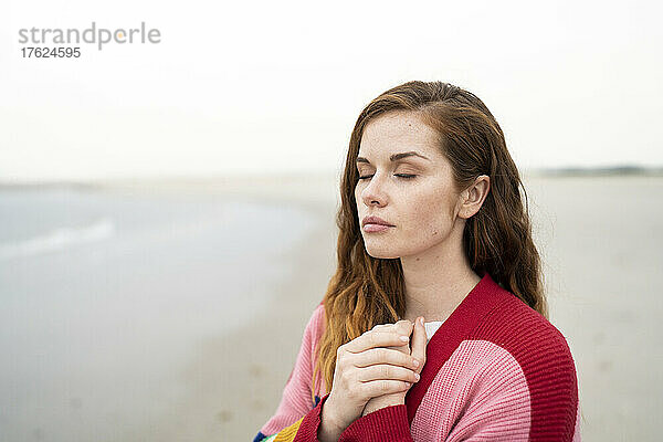 Schöne junge rothaarige Frau steht mit geschlossenen Augen am Strand