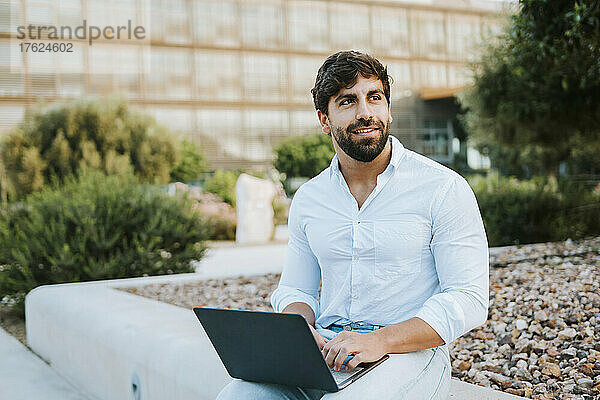 Lächelnder Freiberufler mit Laptop sitzt auf Betonsitz im Büropark