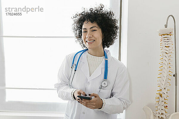 Happy young doctor holding smart phone standing by window at clinic