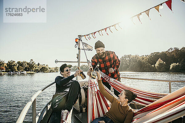 Lächelnde Freunde  die am Bug des Bootes sitzen und Bierflaschen anstoßen