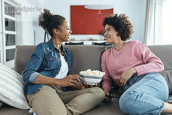 Glückliche Frau hält eine Schüssel Popcorn in der Hand und unterhält sich zu Hause mit einer Freundin auf dem Sofa