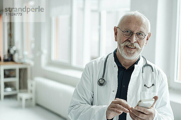 Lächelnder Oberarzt mit Mobiltelefon im Krankenhaus