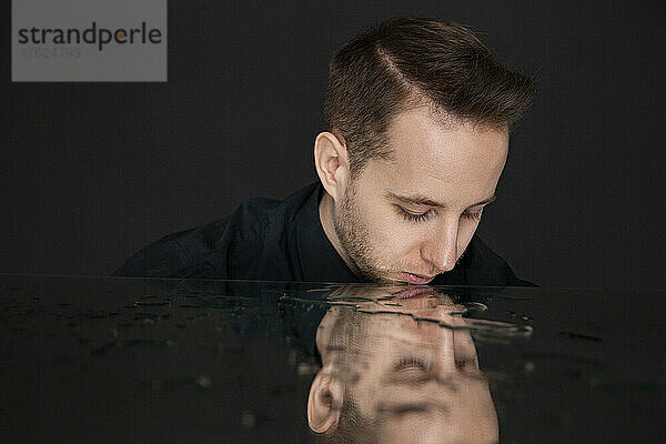 Young man looking at reflection with water on mirror against black background