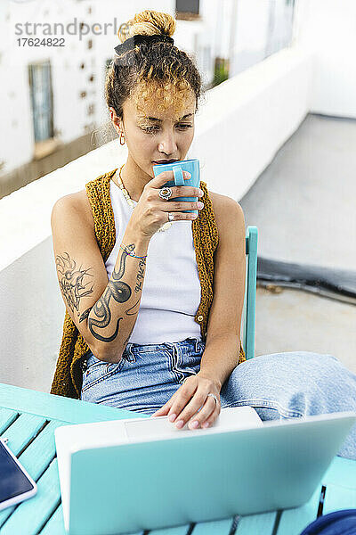 Junge Frau trinkt eine Tasse Tee und sitzt mit Laptop auf dem Dach