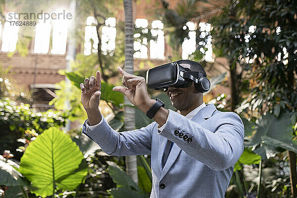 Lächelnder Geschäftsmann mit Virtual-Reality-Simulator gestikuliert mit den Händen vor Pflanzen