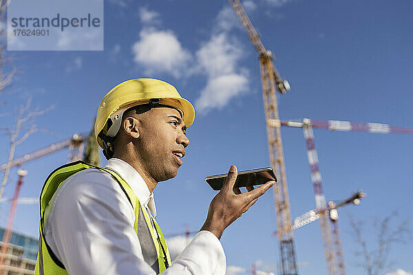 Ingenieur spricht an sonnigem Tag auf der Baustelle mit Smartphone über Lautsprecher