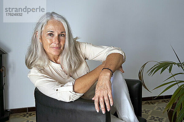 Smiling woman with gray hair sitting on chair in front of wall