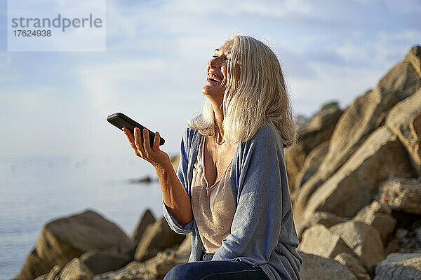 Fröhliche Frau  die an einem sonnigen Tag über den Lautsprecher auf dem Smartphone spricht