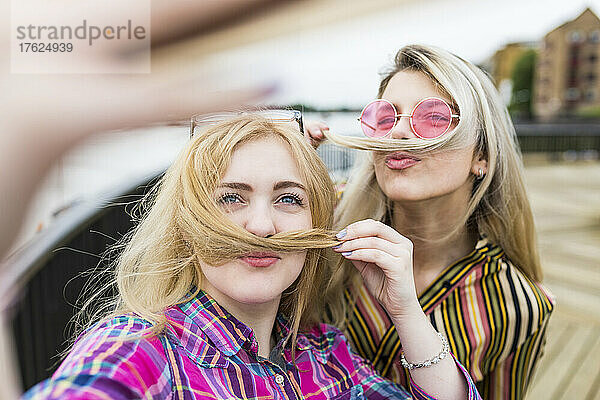 Smiling blond teenagers making mustaches with hair taking selfie