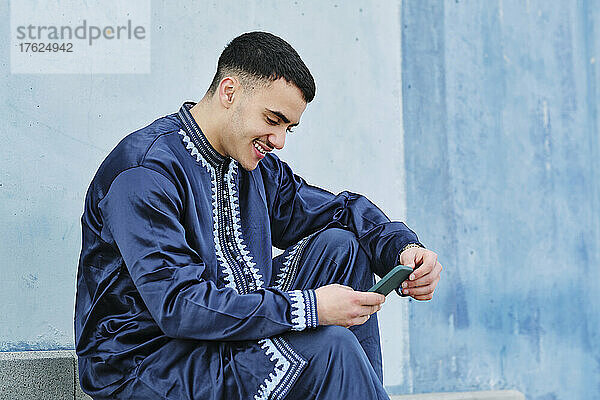 Smiling young man using mobile phone by blue wall