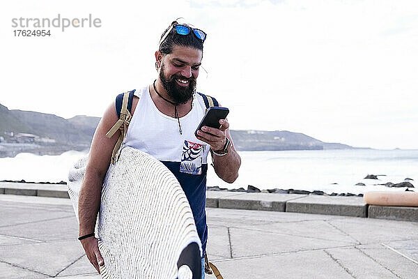Glücklicher Mann mit Surfbrett und Smartphone auf dem Seeweg
