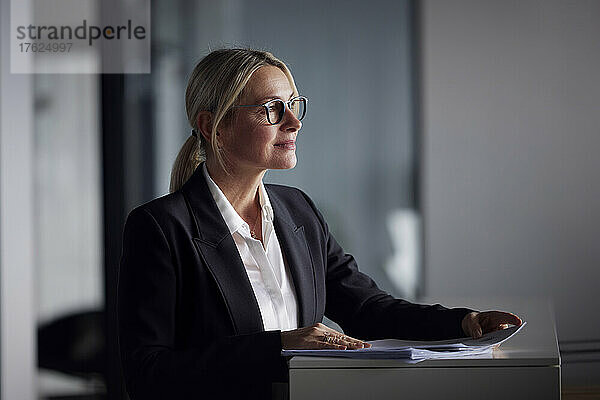 Blonde Geschäftsfrau mit Brille steht mit Dokumenten am Schreibtisch im Büro