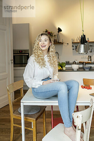 Fröhliche blonde Frau mit Kaffeetasse sitzt zu Hause auf dem Tisch in der Küche