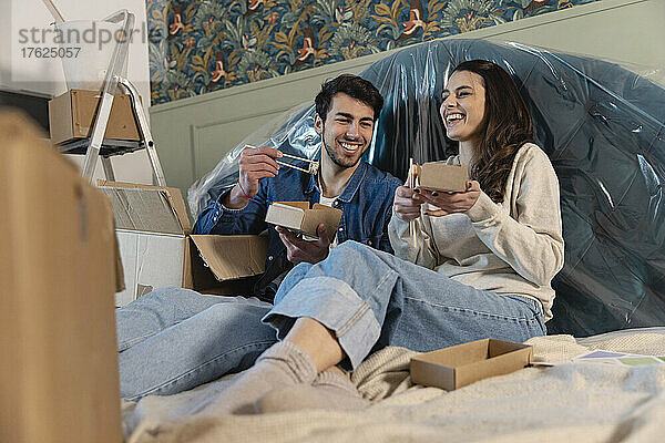 Happy couple having sushi sitting on bed at new home