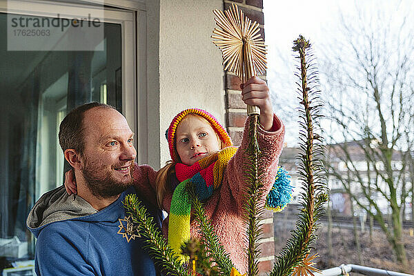 Lächelnder Mann trägt Tochter mit Strickmütze und schmückt den Weihnachtsbaum