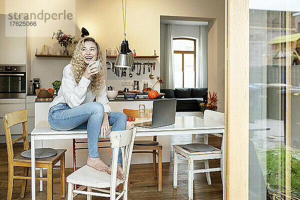 Fröhliche blonde Frau mit Kaffeetasse sitzt auf dem Tisch in der Küche
