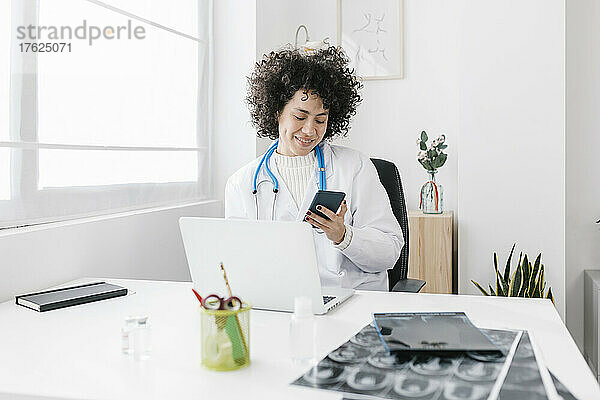 Arzt sitzt mit Laptop und benutzt Mobiltelefon in der medizinischen Klinik