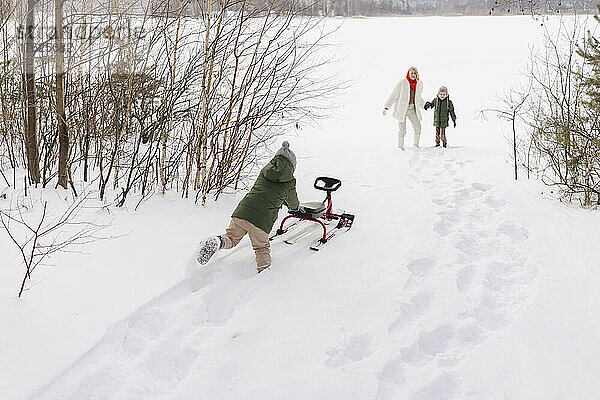 Frau mit Sohn schaut Jungen zu  der im Winter Rodel auf Schnee schiebt