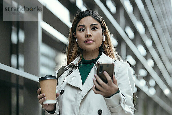 Frau trägt In-Ear-Kopfhörer und hält Smartphone und Einwegkaffeetasse in der Hand