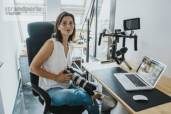 Lächelnder Fotograf mit Digitalkamera sitzt auf einem Stuhl mit Laptop auf dem Tisch im Studio