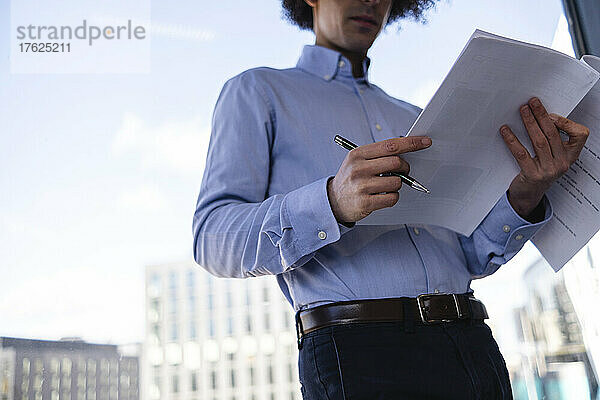Geschäftsmann hält Stift und liest Dokument an Glaswand im Büro gelehnt