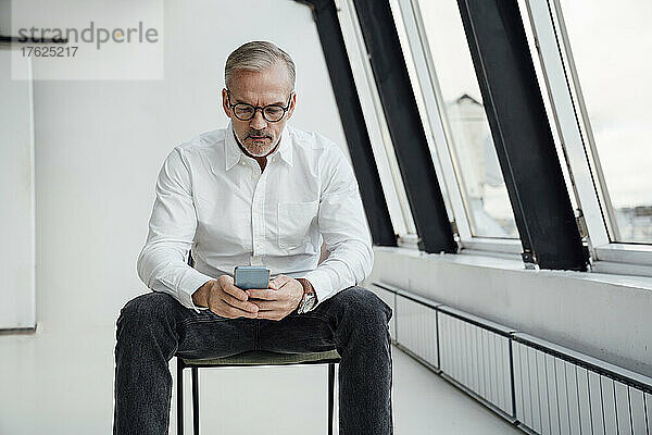 Geschäftsmann mit Brille schreibt Textnachrichten auf dem Mobiltelefon im Büro