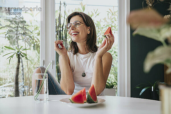 Lächelnde Frau mit Trinkwasser und Wassermelone auf dem Tisch zu Hause