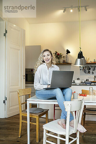 Fröhlicher Freiberufler mit Laptop sitzt zu Hause auf dem Tisch in der Küche