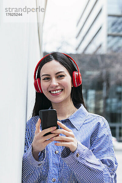 Lächelnde Frau hält ihr Mobiltelefon in der Hand und hört Musik über kabellose Kopfhörer