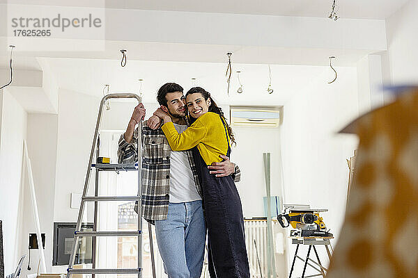 Glückliche Frau umarmt ihren Freund  der an der Leiter im neuen Wohnzimmer steht