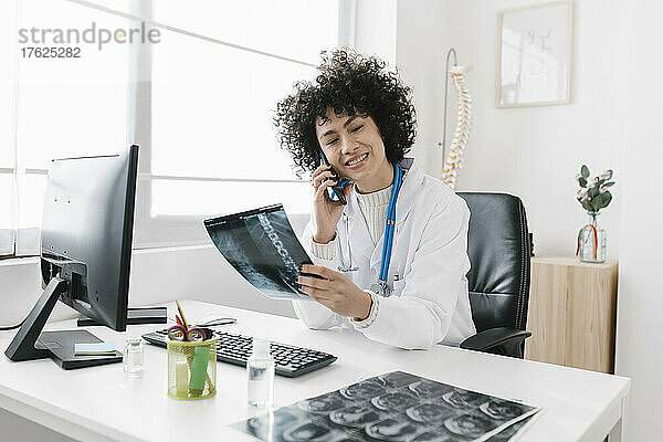 Lächelnder junger Arzt hält Röntgenbild in der Hand und spricht mit dem Mobiltelefon am Schreibtisch im Krankenhaus