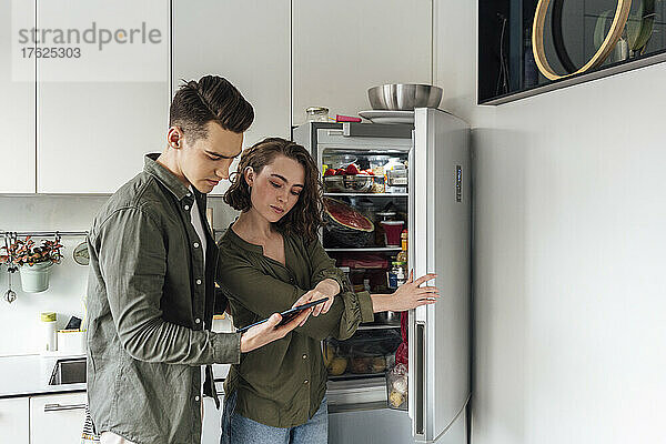 Junger Mann und Frau checken Liste auf Tablet-PC neben Kühlschrank in der Küche
